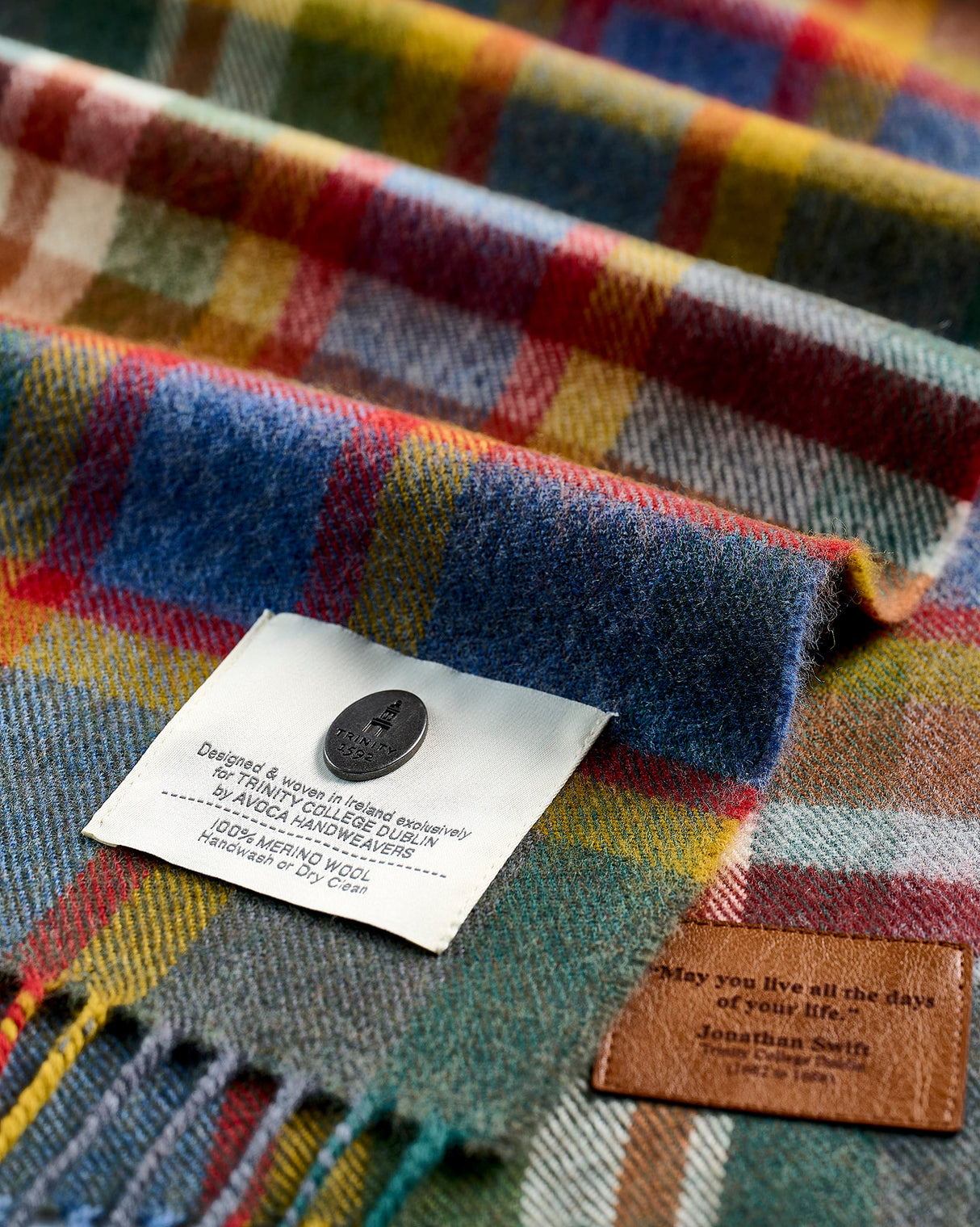 Close-up of a folded plaid scarf or blanket showing its multicolor woven pattern, a white sewn-in label with a black button, and a brown leather brand patch reading 'May you live all the days of your life, Inhabitant 505'.